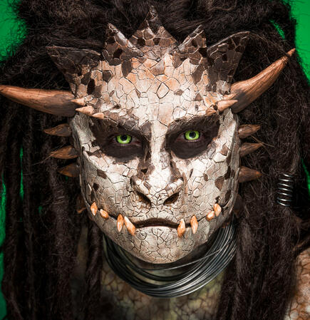 Humanoid reptilian dragon person with brown dreadlocks, pointed horns, yellow eyes, and exposed alligator-like teeth.