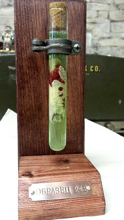 A prop of a slightly mangled, severed human finger in a vial of vibrant green liquid and capped with a cork. A clamp secures it to a wooden mount with a metal nameplate