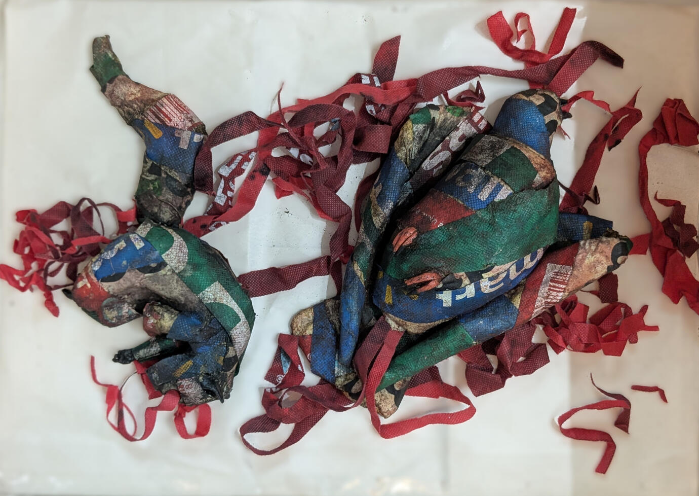 Depicted on a blank background are shapes of a squirrel and a pigeon wrapped in dirty and colourful strips with text on them. Red and crimson strips of fabric with some lettering on them are strewn about chaotically.